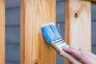 A hand painting a wooden fence plank with a blue paintbrush outdoors. Perfect for DIY and renovation themes.